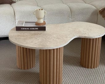 Peut inclure: Une table basse beige en forme de rein avec un plateau en pierre de couleur claire et trois pieds cylindriques en bois. Un livre avec le texte "CATALOG SUBLIME" et un vase crème sont posés sur la table. La table est sur un tapis à motifs.