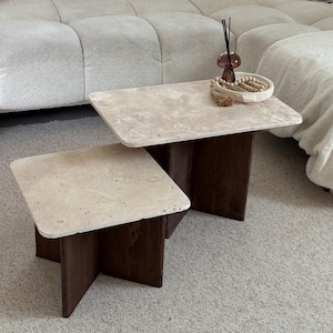 Peut inclure: Ensemble de deux tables basses. Les tables ont des plateaux en pierre beige clair et des bases en bois brun foncé. La plus grande table a un plateau rectangulaire et la plus petite table a un plateau carré. Les tables sont sur un tapis gris clair.