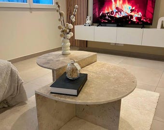 Peut inclure: Une table basse en pierre à deux niveaux avec un plateau arrondi et un livre sur le niveau inférieur. La table est dans un salon avec une cheminée et un vase de fleurs blanches.