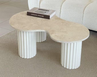 Peut inclure: Une table basse moderne à la forme organique unique. Le plateau est de couleur beige clair, et la base est composée de trois pieds cylindriques cannelés blancs. Un livre avec le texte "CATALOG SUBLIME" est posé sur la table.