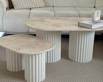 Peut inclure: Deux tables basses ovales. Les plateaux sont beiges clairs et texturés. Les bases sont blanches, cylindriques et cannelées. Les tables sont devant un canapé blanc avec des coussins beiges. Le sol est recouvert d'une moquette beige.