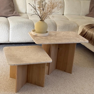 Peut inclure: Deux tables basses carrées avec des plateaux en pierre beige clair et des bases en bois brun clair. Les tables sont de tailles différentes et sont placées côte à côte. Un vase beige avec des fleurs séchées est posé sur la plus grande table. Les tables sont dans un salon.