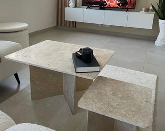 Peut inclure: Une table basse en travertin beige avec une table d'appoint assortie. Un livre et une tasse noirs sont posés sur la table basse. Les tables ont un plateau rectangulaire et un socle plein. Les tables sont dans un salon.