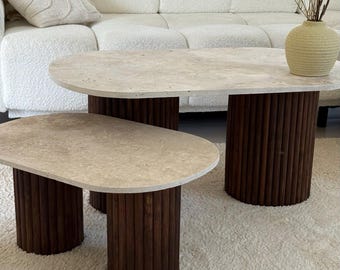 Peut inclure: Deux tables basses ovales avec des plateaux en pierre claire et des bases cylindriques en bois brun foncé. Les tables sont de tailles différentes et reposent sur un tapis blanc. Un canapé blanc et un vase décoratif sont en arrière-plan.