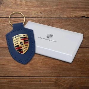 May include: A blue leather keychain with a gold ring and a gold and red Porsche emblem. The keychain is next to a white box with the Porsche logo and text. The background is a wooden surface.