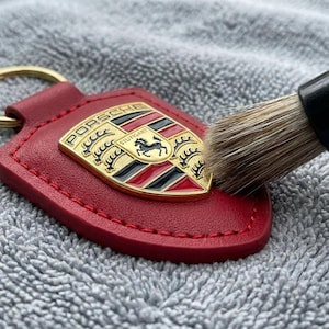 May include: Red leather keychain with a gold Porsche emblem. The emblem features the Porsche crest with a black horse, red and black stripes, and the words "Porsche" and "Stuttgart". A cleaning brush is touching the emblem.