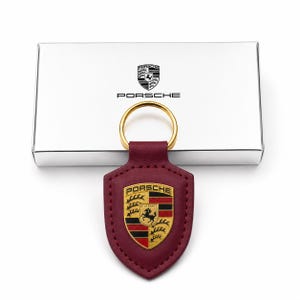 May include: A burgundy leather keychain featuring a shield-shaped emblem with the Porsche logo in gold, black, and red. The keychain includes a gold-toned metal ring and is positioned in front of a silver rectangular box with the Porsche logo.