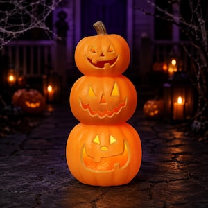 May include: A vertical stack of three orange jack-o'-lantern pumpkins with carved faces. Each pumpkin features cutouts for triangular eyes, a nose, and a wide, jagged mouth. The pumpkins are illuminated from within, casting a warm, bright glow.