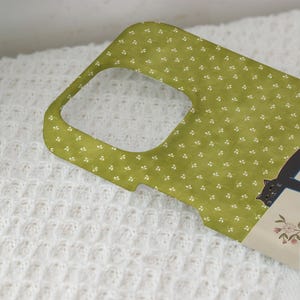 May include: A green phone case with a repeating white floral pattern. A black cat illustration is visible on the side. The case is designed to fit a phone, with cutouts for the camera and buttons.