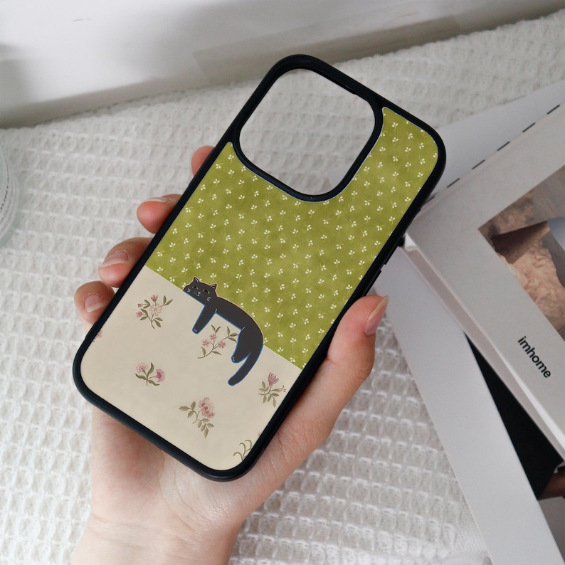 May include: Black phone case with a black border. The case features a two-tone design: a green upper section with white floral patterns and a beige lower section with a black cat illustration and pink floral accents.