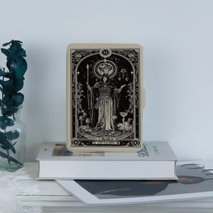 May include: A beige tarot card case featuring a black and white illustration of a robed figure with a staff, surrounded by celestial elements. The text "THE MAGINN" is at the bottom. The case is resting on a stack of books.