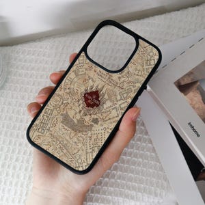 Può includere: Custodia per telefono nera con un design a mappa, che include le parole "The Marauders Map" in un emblema rosso. La custodia ha un bordo nero e un ritaglio per la fotocamera. Il design della mappa è nei toni del marrone e beige.
