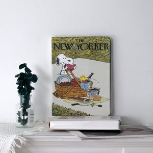 Könnte beinhalten: Ein Buch mit einem Cover mit einer Snoopy-Illustration aus The New Yorker. Das Bild zeigt Snoopy bei einem Picknick mit einem Korb voller Essen und einer Flasche Wein. Das Buch liegt auf einer weißen Oberfläche.