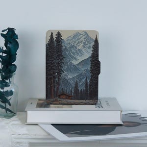 May include: A rectangular e-reader case featuring a printed illustration of a mountain range with snow-capped peaks, a dense forest of tall pine trees, and a small wooden cabin nestled at the base of the mountains in shades of blue and brown.