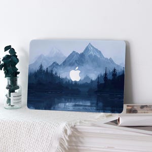 May include: A laptop case featuring a scenic mountain landscape in shades of blue and white. The design includes a lake, trees, and a mountain range with a white Apple logo. The case is on a white surface.