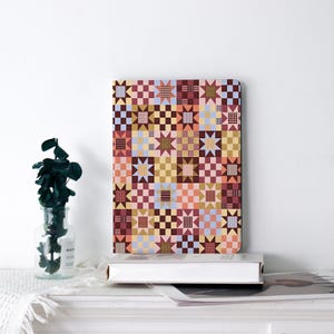 May include: A rectangular notebook featuring a repeating quilt-inspired pattern of stars and checkered squares in shades of burgundy, mustard yellow, light blue, and soft pink. The cover design consists of a grid of geometric shapes.