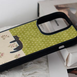 May include: Black phone case with a whimsical design. The case features a black cat illustration on a cream-colored background with pink floral accents. A section of the case has a green background with a white floral pattern.