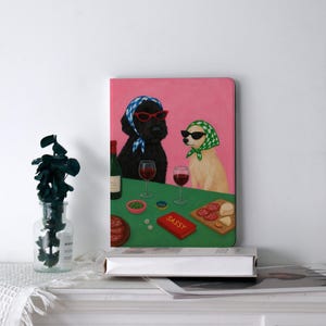 May include: A pink notebook featuring a whimsical illustration of two dogs wearing sunglasses and headscarves, enjoying wine and snacks. A red book on the table reads "Sassy". A bottle of wine and glasses are also on the table.