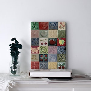 May include: A decorative wall art piece composed of 25 square ceramic tiles. Each tile features a unique embossed design, including a heart, snail, butterfly, and cherries. The tiles are arranged in a grid pattern, showcasing a variety of colors and textures.
