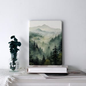 May include: A landscape art print featuring a misty forest scene with tall evergreen trees and rolling hills. The artwork is in shades of green and gray, creating a serene and natural aesthetic. The print is displayed on a white surface.