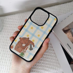 May include: Black phone case with a whimsical design. The case features a brown bear pouring coffee, set against a light blue plaid background with yellow stars. The case has a black border.