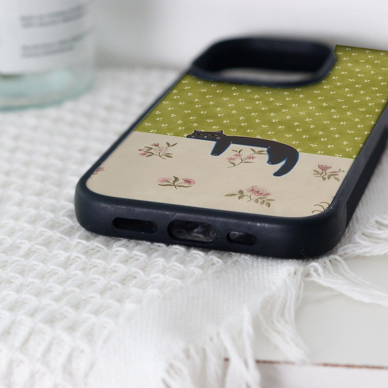 May include: A black phone case featuring a whimsical design. The case is divided into two sections: a green upper half with a white floral pattern and a lower half with a beige floral pattern. A black cat is depicted lounging on the beige section.