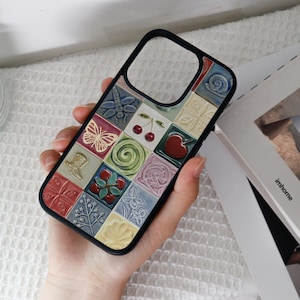 May include: Black phone case with a colorful, textured tile design. The tiles feature various embossed images, including a butterfly, cherries, a heart, and floral patterns. The case has a black border and a cutout for the camera.