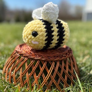 May include: A crocheted bee plush toy with yellow and black stripes, white wings, and a cute face. The bee is sitting on a brown wicker basket.