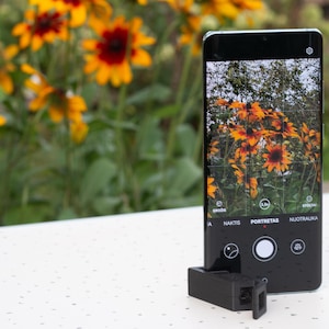 Puede incluir: Un teléfono inteligente negro con un accesorio de cámara se encuentra sobre una superficie blanca, mostrando una foto de flores amarillas y rojas. El fondo presenta más flores similares.