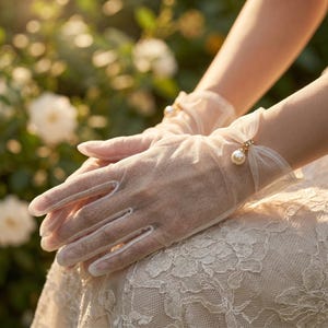 May include: Sheer, ivory-colored gloves with delicate pearl accents. The gloves are fingerless and adorned with small, dangling pearls at the wrist. The background features soft, blurred greenery and sunlight, creating a romantic and elegant aesthetic.