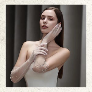 Könnte beinhalten: Transparente, weiße Handschuhe mit kleinen Perlen verziert. Die Handschuhe reichen bis zum Oberarm und bestehen aus einem zarten, durchsichtigen Stoff. Die Perlen sind um den Ellbogen angeordnet und verleihen Eleganz.