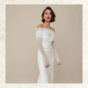 May include: A white, off-the-shoulder wedding dress with sheer, long sleeves embellished with small pearls. The dress features a fitted bodice and a flowing skirt. The model has short, dark hair and red lipstick.