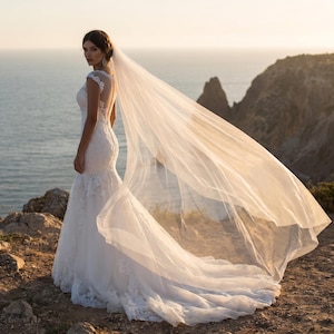 May include: A white, lace wedding dress with a mermaid silhouette. The dress features cap sleeves and a sheer back with button detailing. A long, flowing veil billows in the wind. The scene is set outdoors with a coastal backdrop.