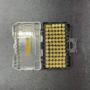 May include: A clear plastic and black ammunition case, open to reveal rows of brass 9mm bullets. The case is labeled "9MM" in yellow and has the "DEWALT" logo. The case is on a dark gray surface.