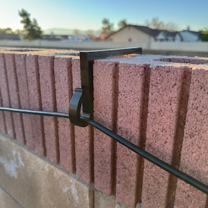 May include: A black metal bracket is attached to a red brick wall, holding a black wire. The bracket is designed to secure the wire, likely for outdoor lighting or electrical purposes. The background shows a clear blue sky and residential buildings.