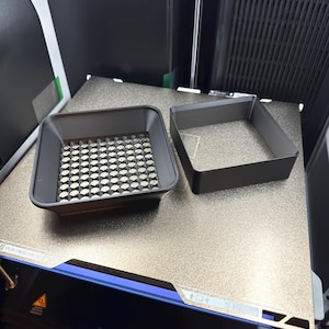 May include: Two dark gray, square-shaped 3D-printed objects sit on a metallic surface. One object has a patterned interior, while the other is a simple frame. The objects are likely components for a 3D printer, showcasing a modern manufacturing process.