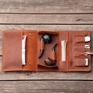 May include: A brown leather tobacco pipe case, open to reveal a pipe, pipe tools, and accessories. The case is made of brown leather and has multiple compartments. The background is a wooden surface.