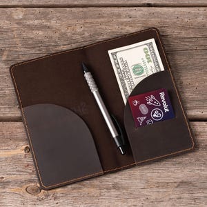 May include: A dark brown leather checkbook cover with contrast stitching, featuring two interior pockets. One pocket holds a one hundred dollar bill, and the other contains a purple Revolut card with white icons. A silver pen rests in the center.