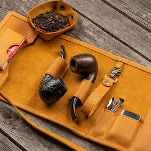 May include: A tan leather tobacco pipe roll featuring two wooden pipes, a small detachable tobacco tray, a metal lighter, pipe cleaning tools, and white pipe cleaners. The roll includes dedicated slots for each item and a strap closure.