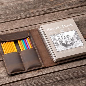 Op de afbeelding: Open leren schetsboek met spiraalbinding en de tekst "Sketch Book" op de kaft. Het schetsboek is open op een pagina met een tekening van een gondel. Een leren etui met gele en gekleurde potloden ligt naast het schetsboek.