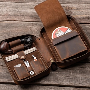 May include: A brown leather tobacco pipe case featuring a zippered closure, an interior compartment for a pipe, a dedicated slot for pipe cleaners, and multiple pockets for accessories. A round tobacco tin is stored in the upper section.