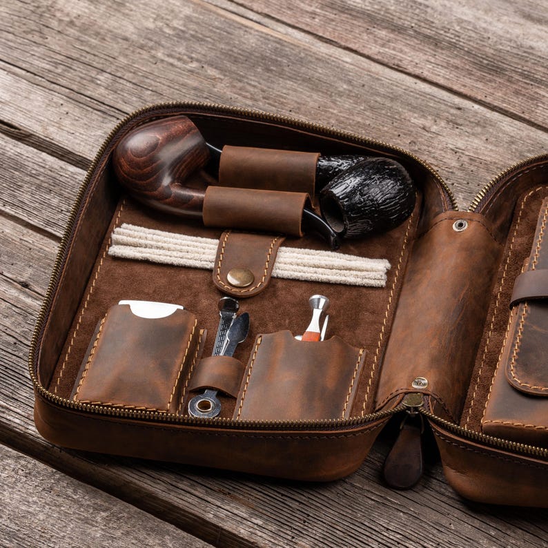 May include: A brown leather zippered case containing two tobacco pipes, a set of white pipe cleaners, a metal pipe tool, and a small metal lighter. The interior features dedicated leather slots and loops to secure each item in place.