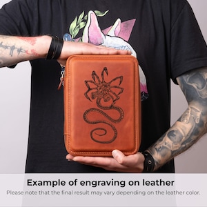 K&ouml;nnte beinhalten: Ein rechteckiges Etui aus hellbraunem Leder mit einer detaillierten, dunkel lasergravierten Illustration eines Facehugger-Wesens auf der Vorderseite. Das Etui hat abgerundete Ecken, einen festen Rahmen und einen sichtbaren Rei&szlig;verschluss an der Seite.