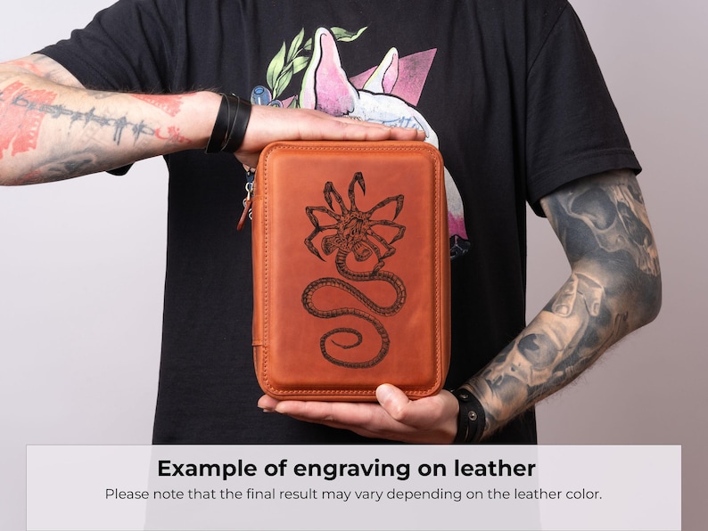 K&ouml;nnte beinhalten: Ein rechteckiges Etui aus hellbraunem Leder mit einer detaillierten, dunkel lasergravierten Illustration eines Facehugger-Wesens auf der Vorderseite. Das Etui hat abgerundete Ecken, einen festen Rahmen und einen sichtbaren Rei&szlig;verschluss an der Seite.