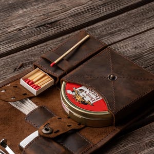 K&ouml;nnte beinhalten: Eine dunkelbraune Tabaktasche aus Leder mit einem speziellen Fach f&uuml;r eine runde Metalldose mit der Aufschrift Devil's Holiday, einem kleinen Schlitz f&uuml;r eine Streichholzschachtel mit roten K&ouml;pfen und weiteren F&auml;chern f&uuml;r Zubeh&ouml;r mit N&auml;hten.