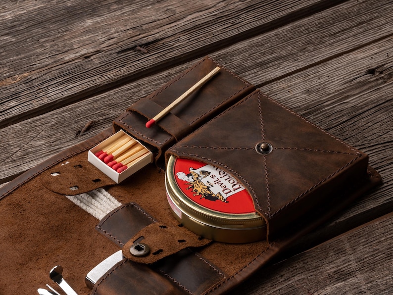 K&ouml;nnte beinhalten: Eine dunkelbraune Tabaktasche aus Leder mit einem speziellen Fach f&uuml;r eine runde Metalldose mit der Aufschrift Devil's Holiday, einem kleinen Schlitz f&uuml;r eine Streichholzschachtel mit roten K&ouml;pfen und weiteren F&auml;chern f&uuml;r Zubeh&ouml;r mit N&auml;hten.