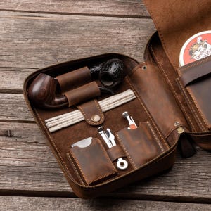 May include: A brown leather tobacco pipe case featuring a zippered closure, containing a dark wood pipe, a textured black pipe, white pipe cleaners, and metal pipe tools stored in dedicated leather slots, with a round tobacco tin visible inside.