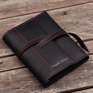 May include: Black leather journal with red stitching and a strap closure. The journal is embossed with the words "Death Note".