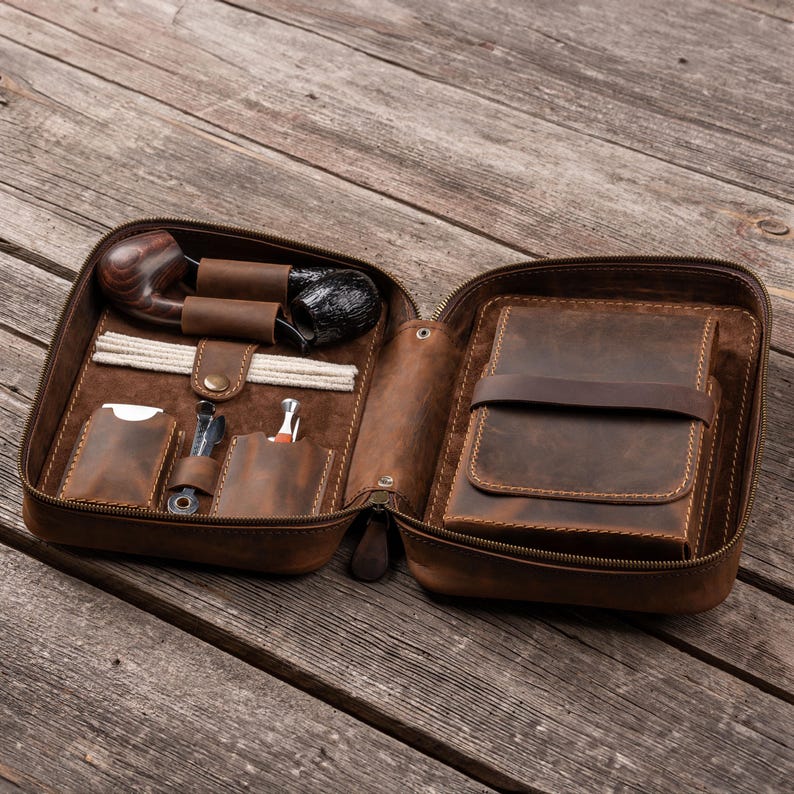 May include: A brown leather zippered case containing a dark wood tobacco pipe, white pipe cleaners, a metal pipe tool, and a small metal lighter. The interior features dedicated leather slots and a large pouch secured by a horizontal leather strap.