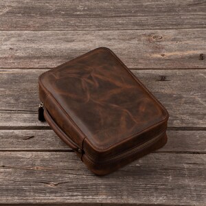 May include: A rectangular brown leather travel case with a smooth, distressed finish and a wrap-around zipper closure. The case features a sturdy side handle and rounded corners, showcasing a deep, rich brown color with natural variations.